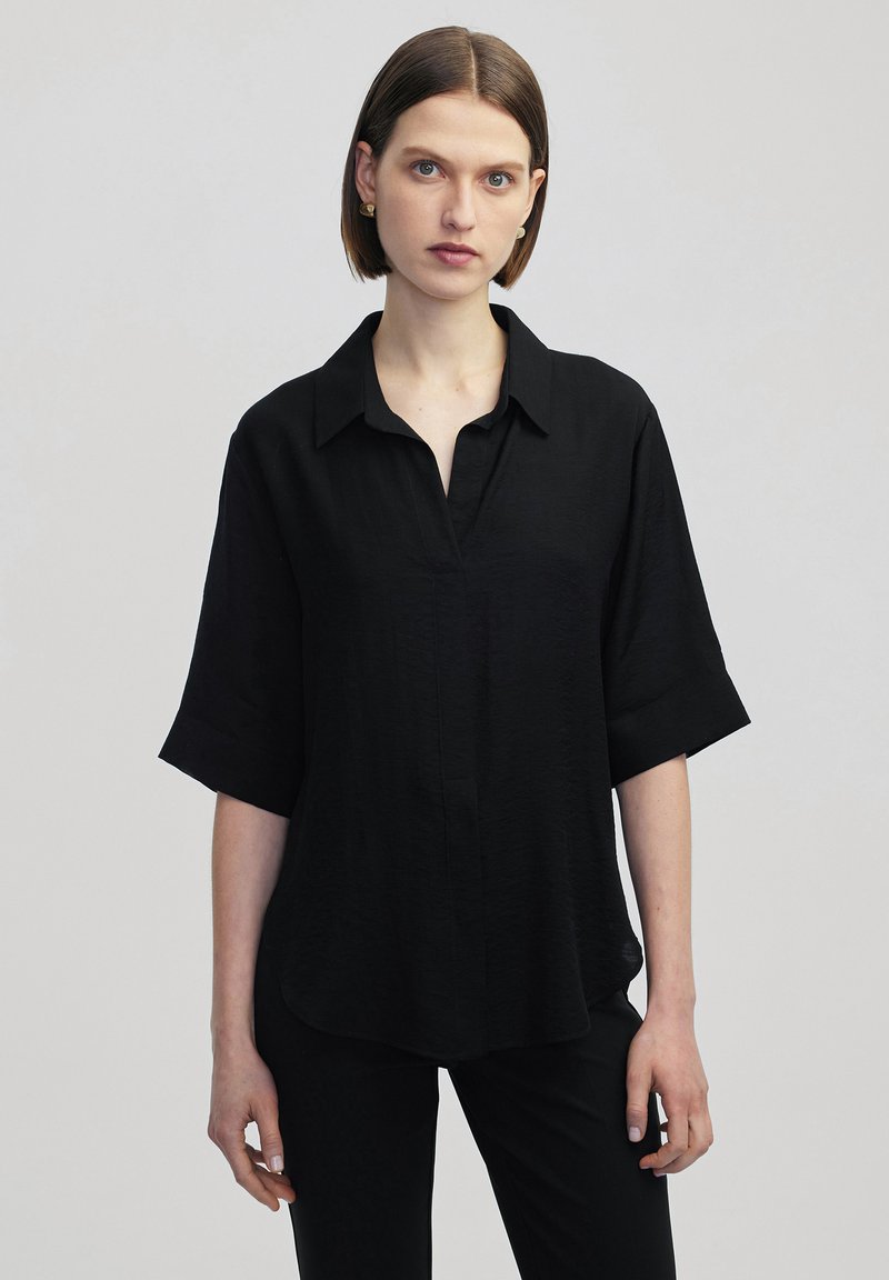 Blusa nera a maniche corte con colletto, caratterizzata da una vestibilità comoda e un tessuto leggermente texturizzato. Immortalata su uno sfondo chiaro.