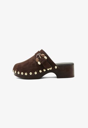 Bruine suède klomp met houten plateauhak, decoratieve gouden studs langs de zool en een bandje met ronde steenversieringen.