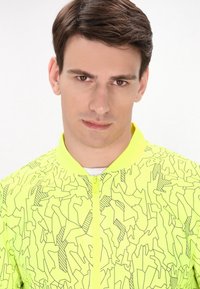 Jachetă galben neon cu un model geometric, având un guler rotund și un fermoar frontal. Material ușor cu o textură netedă.