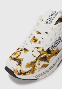 White sneaker with gold baroque pattern, white laces, and Versace Jeans Couture logo on tongue, side, and sole.