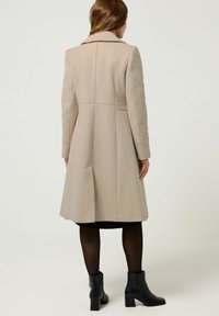 Beige wollen jas met een getailleerde silhoeët en brede kraag, voorzien van achternaden en een uitlopende zoom. Draag het over zwarte panty’s en enkellaarsjes.
