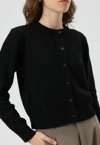 Zwarte gebreide cardigan met een ronde hals, lange mouwen en knoopsluiting. Geribbelde boorden en zoom. Zachte textuur, minimalistisch ontwerp.