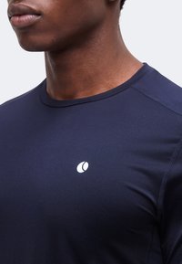 Άνδρας που φοράει ναυτικό μπλε αθλητικό πουκάμισο με μικρό λευκό κυκλικό λογότυπο στο αριστερό στήθος, εμφανίζεται από τον λαιμό μέχρι το μέσο του κορμού.