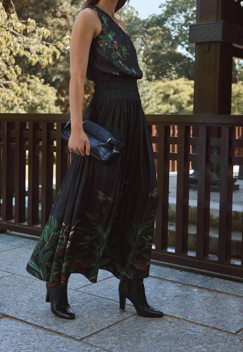 Femme portant une robe noire sans manches à motifs floraux et des bottines noires à talons, tenant une pochette noire en se tenant à l'extérieur.