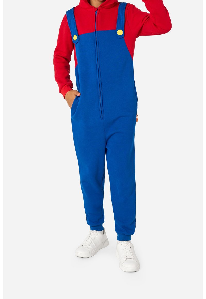Enfant portant une salopette bleue avec des boutons jaunes sur un sweat à capuche rouge et des baskets blanches, debout avec une main dans la poche et l'autre levée.