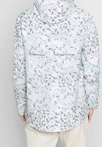 Lätt jacka i en ljusgrå färg med ett spritt abstrakt mönster i mörkgrå och gröna nyanser. Har en huva och bakfickor.