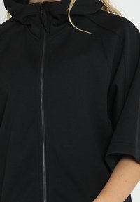 Černá mikina s kapucí a plným zipem, volný střih, měkká textura a krátké rukávy. Vyznačuje se vysokým límcem a minimálními švy.