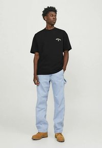 Black crew-neck t-shirt with graphic detailing on the chest, paired with light blue jeans and tan work boots.
