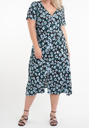 Femme portant une robe midi noire à motif floral bleu et blanc et des sandales noires à brides, debout, les mains lâches sur les côtés.