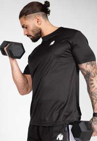 Black short-sleeved athletic shirt with a small white logo, worn with black shorts, holding hexagonal dumbbells. Smooth texture, fitted design.