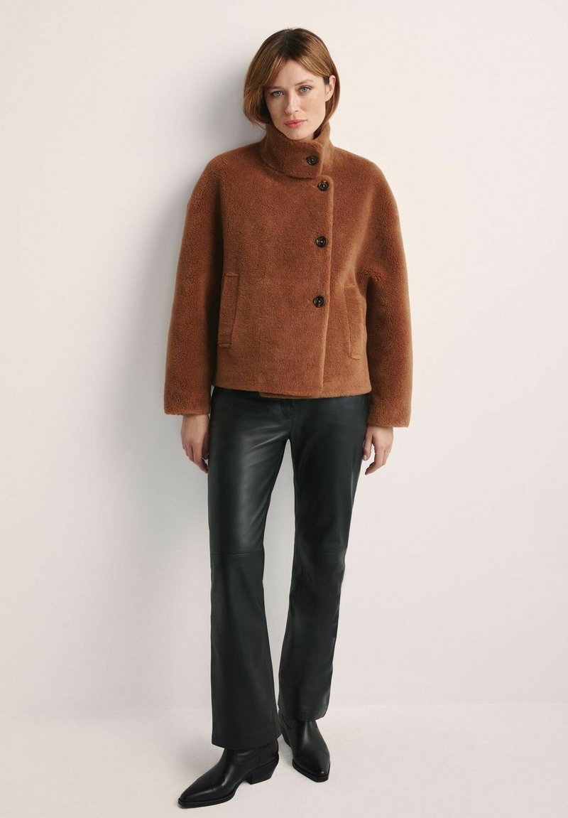 Brązowa kurtka o futrzastej teksturze z wysokim kołnierzem, trzema guzikami i kieszeniami z przodu; zestawiona z czarnymi skórzanymi spodniami i botkami za kostkę.