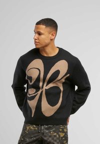 Schwarzer Strickpullover mit einem großen beigen Schmetterlingsdesign auf der Vorderseite, runder Ausschnitt und langen Ärmeln. Glatte Textur.