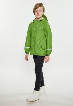 Groene waterdichte regenjas met een capuchon, drukknopen en rits, witte reflecterende strepen en zijzakken. Draagt over zwarte broeken en witte sneakers.