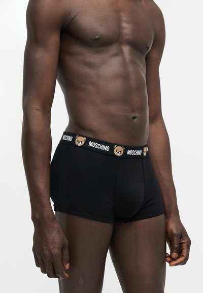 Ανδρικός κορμός που φοράει μαύρα μποξεράκια Moschino με ελαστική μέση διακοσμημένη με πρόσωπα αρκουδάκια και επαναλαμβανόμενο λογότυπο Moschino.