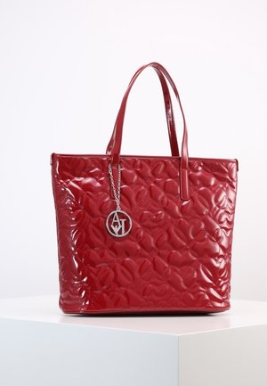 Sac à main matelassé rouge brillant avec deux anses et un pendentif circulaire argenté suspendu à une anse, exposé sur un socle blanc.