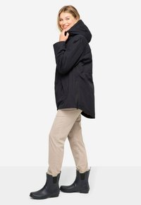 Chaqueta impermeable negra con capucha, combinada con pantalones beige claros y botas de tobillo negras con paneles elásticos laterales. Cuenta con bolsillos laterales.
