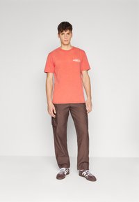 Camiseta de algodón roja con mangas cortas y logotipo en el pecho izquierdo, combinada con pantalones cargo marrones que tienen bolsillos laterales. Zapatillas blancas.