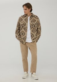 Brązowa koszula w geometryczne wzory, biała koszulka pod spodem, beżowe spodnie sztruksowe i sneakersy. Gładki materiał i luźny krój.