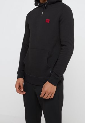Mężczyzna w czarnej bluzie z kapturem, z przodem z kieszenią typu kangurek oraz małym czerwonym kwadratowym logo na klatce piersiowej, w zestawie z czarnymi spodniami.