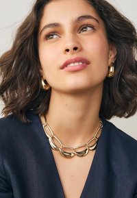Frau mit kurzen, welligen Haaren, die eine goldene, grobe Kettenhalskette und passende goldene Creolen trägt, gekleidet in ein dunkles Oberteil und leicht nach oben schaut.
