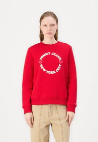 Rød hettegenser laget av bomull, med et rundt hvitt grafisk trykk som står "TOMMY JEANS NEW YORK CITY" i midten. Klassisk passform og ribbestrikkede mansjetter.