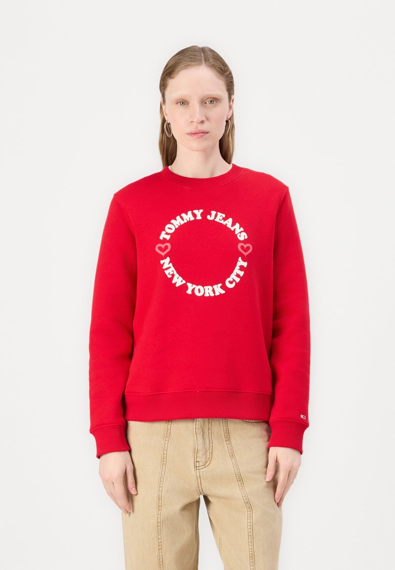 Rød hettegenser laget av bomull, med et rundt hvitt grafisk trykk som står "TOMMY JEANS NEW YORK CITY" i midten. Klassisk passform og ribbestrikkede mansjetter.