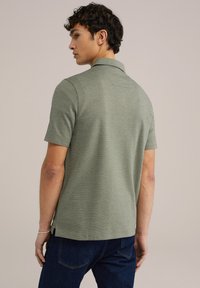 Tricou polo verde cu manșon scurt, cu o textură țesută, guler colorat și fante laterale. Purtați parțial întors, evidențiind detaliile din spate.
