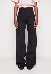Pantalones negros de pierna ancha hechos de tejido texturizado, con una cintura alta, bolsillos frontales y cierre de botón, combinados con zapatos negros de suela gruesa.