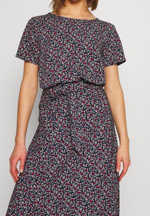 Femme portant une robe à manches courtes bleu marine avec un petit motif floral rouge et blanc et une ceinture en tissu nouée à la taille.