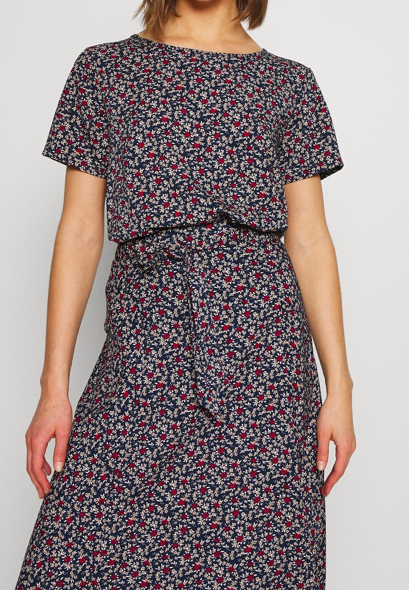Woman wearing navy blue short-sleeve dress with red and white small floral pattern and fabric belt tied at waist.