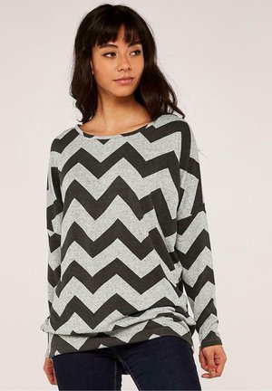 Kvinde iført en løs grå sweater med brede sorte zigzagstriber, kombineret med mørke jeans, står mod en ensfarvet baggrund.
