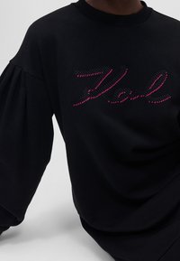 Sweat-shirt noir avec encolure ronde, manches bouffantes et coutures décoratives roses formant le mot "Karl" sur le devant.