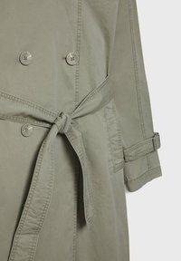 Vaaleanoliivinvihreä trench-takki, jossa on kaksiriviset napit, sidottava vyö ja säädettävät hihansuut, valmistettu kestävästä puuvillakankaasta.