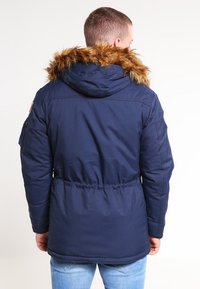 Mornarsko modra parka jakna z kapuco obloženo s krzneno podlogo, elastičnim pasom in stranskimi žepi. Gladek material s protokolarno teksturo, minimalne oznake blagovne znamke.