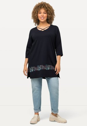 Tunic blu navy con scollatura incrociata, orlo decorato con motivi floreali colorati, abbinata a jeans chiari e scarpe beige.