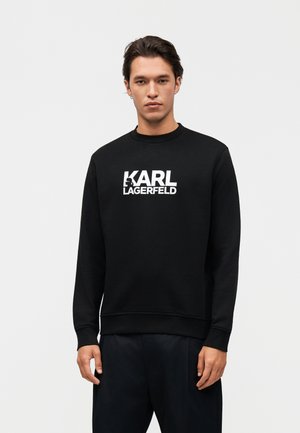 Jonge man die een zwart sweatshirt draagt met de tekst "KARL LAGERFELD" in het wit, staand tegen een effen lichte achtergrond.