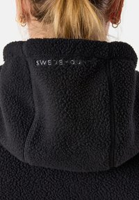 Schwarze Fleece-Hoodie-Jacke aus strukturiertem Stoff. Sie verfügt über ein sichtbares Logo, das auf dem Rücken gestickt ist. Einfaches Design mit lockerer Passform.