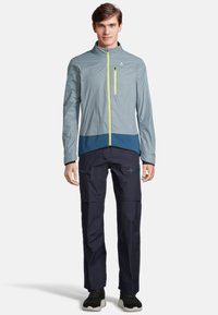 Coupe-vent léger, gris, avec une section inférieure bleu marine et une fermeture éclair jaune vif. Associé à un pantalon bleu marine avec des poches zippées.