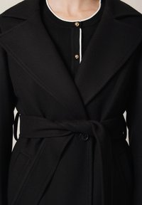 Czarny płaszcz zapinany, wykonany z miękkiego materiału, z szerokim kołnierzem, długimi rękawami i przewiązywanym paskiem w talii. Zapięcie na guziki z przodu i kieszenie po bokach.