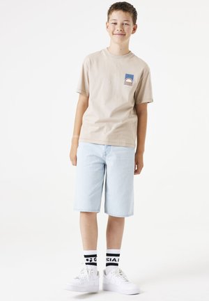 T-shirt en coton beige avec un petit graphique bleu et rouge, short en denim bleu clair, et baskets blanches avec des chaussettes à accents noirs.