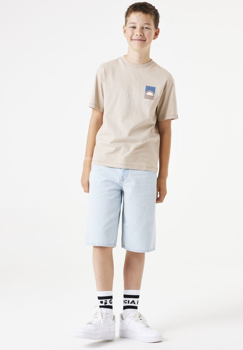 T-shirt en coton beige avec un petit graphique bleu et rouge, short en denim bleu clair, et baskets blanches avec des chaussettes à accents noirs.