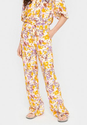 Kobieta ubrana w luźne spodnie w kwiatowy wzór i dopasowany top w odcieniach żółci, fioletu i brązu, stoi z jedną ręką w kieszeni, ma na sobie sandały.