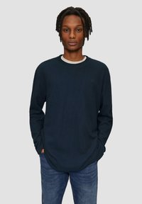 Donkerblauwe longsleeve shirt met ribbelstructuur en ronde hals, gelaagd over een wit T-shirt, gecombineerd met denim jeans.