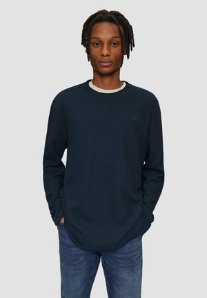 Chemise à manches longues bleu foncé avec texture côtelée et col rond, superposée à un t-shirt blanc, assortie à un jean en denim.