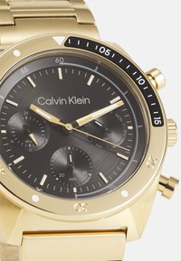 Calvin Klein gold-tone watch with a black textured dial, three subdials, gold hour markers, and a metal bracelet strap.