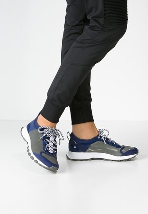 Person som bär svarta joggingbyxor och blå-grå sneakers med vita mönstrade snören, står med ena foten över den andra.
