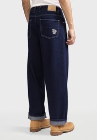 Mann trägt dunkelblaue weite Jeans mit einem Logo auf der Gesäßtasche und beigen Schnürstiefeln, steht vor einem einfarbigen Hintergrund.