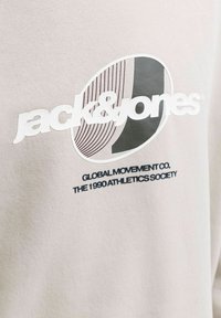 Logo Jack & Jones avec des graphismes rayés et unis sur un tissu clair, texte en dessous : Global Movement Co. The 1990 Athletics Society.