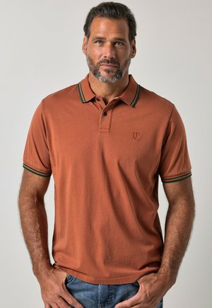 Mann mit kurzem Bart, der ein rostfarbenes Poloshirt mit grün gestreiftem Kragen und Ärmeln trägt, steht vor einem einfachen hellen Hintergrund.