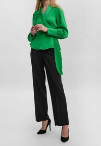 Blusa verde in satin avvolgente con maniche lunghe e design drappeggiato, abbinata a pantaloni neri a vita alta e tacchi a spillo neri.